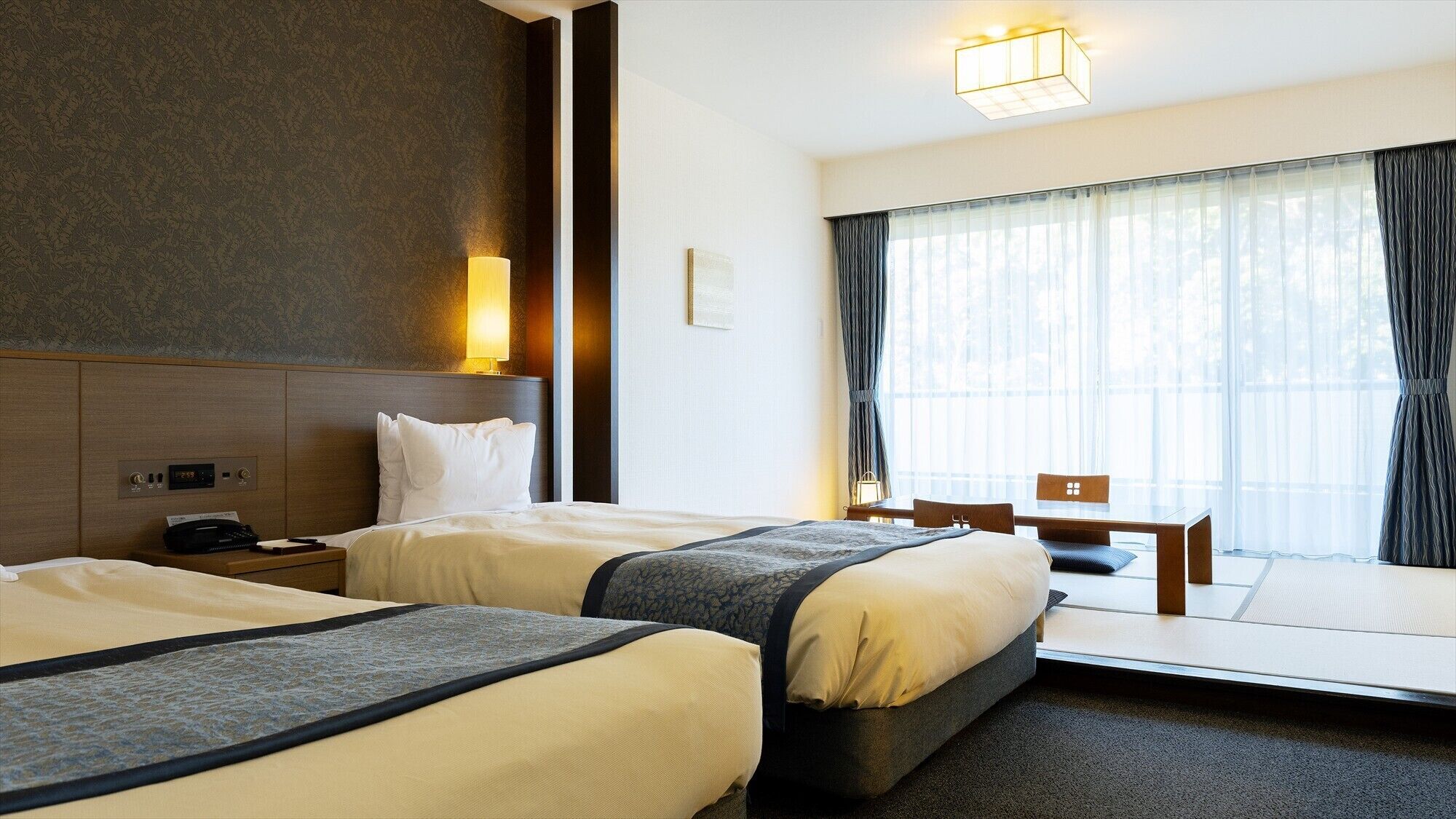 Habitación de hotel con dos camas, un escritorio, una silla, un ventanal con cortinas, una lámpara de noche y un panel de control en la pared.