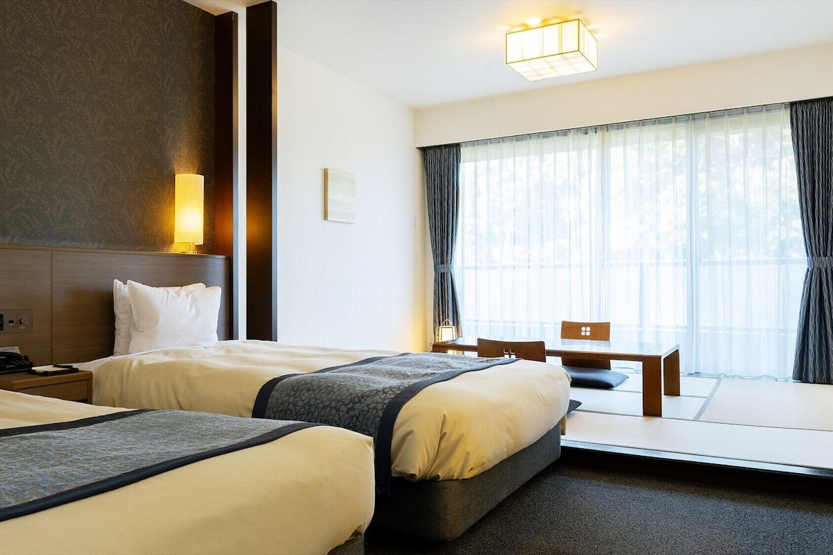 Habitación de hotel con dos camas, un escritorio, una silla, un ventanal con cortinas, una lámpara de noche y un panel de control en la pared.