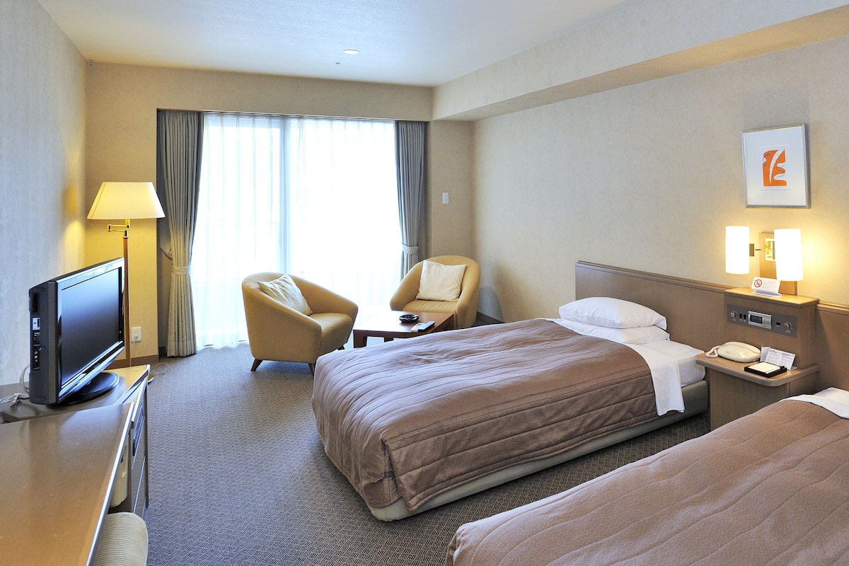 Una habitación de hotel con dos camas, un escritorio con televisor, una silla y una ventana con cortinas.