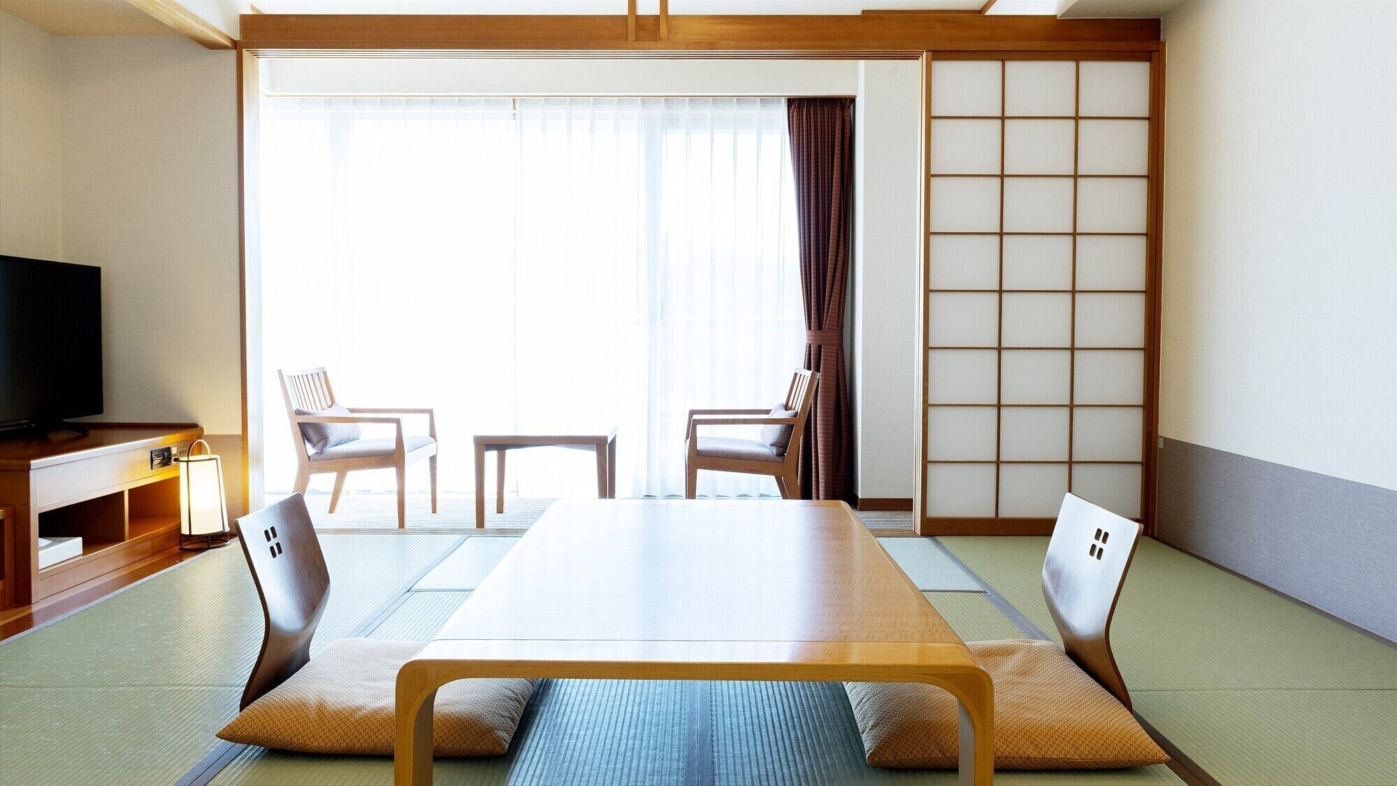 Una habitación japonesa tradicional con suelo de tatami, una mesa baja, dos sillas, un televisor y un panel corredero shoji.