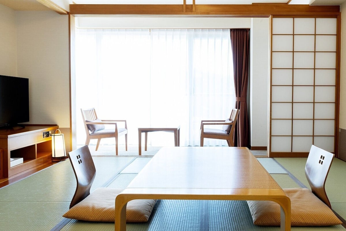 Una habitación japonesa tradicional con suelo de tatami, una mesa baja, dos sillas, un televisor y un panel corredero shoji.