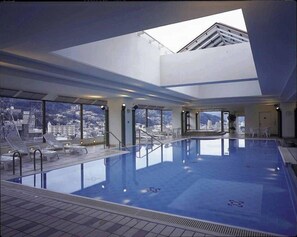 Una amplia piscina cubierta con vistas a la ciudad, tragaluces y una terraza con sillones de descanso.