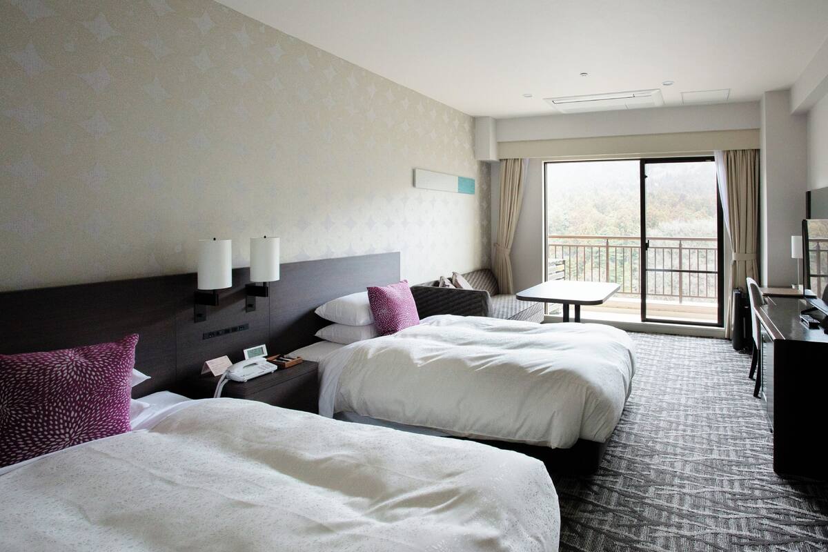 Ein Hotelzimmer mit zwei Betten, einem Schreibtisch, einem Stuhl, einem kleinen Tisch und einem Balkon mit Blick.