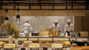 Une cuisine moderne où des chefs préparent des plats, avec des meubles en bois, et divers ustensiles de cuisine et ingrédients disposés sur le plan de travail.