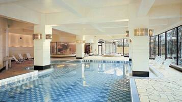 A large indoor swimming pool with clear blue tiles, surrounded by lounge chairs and a stone-tiled floor.