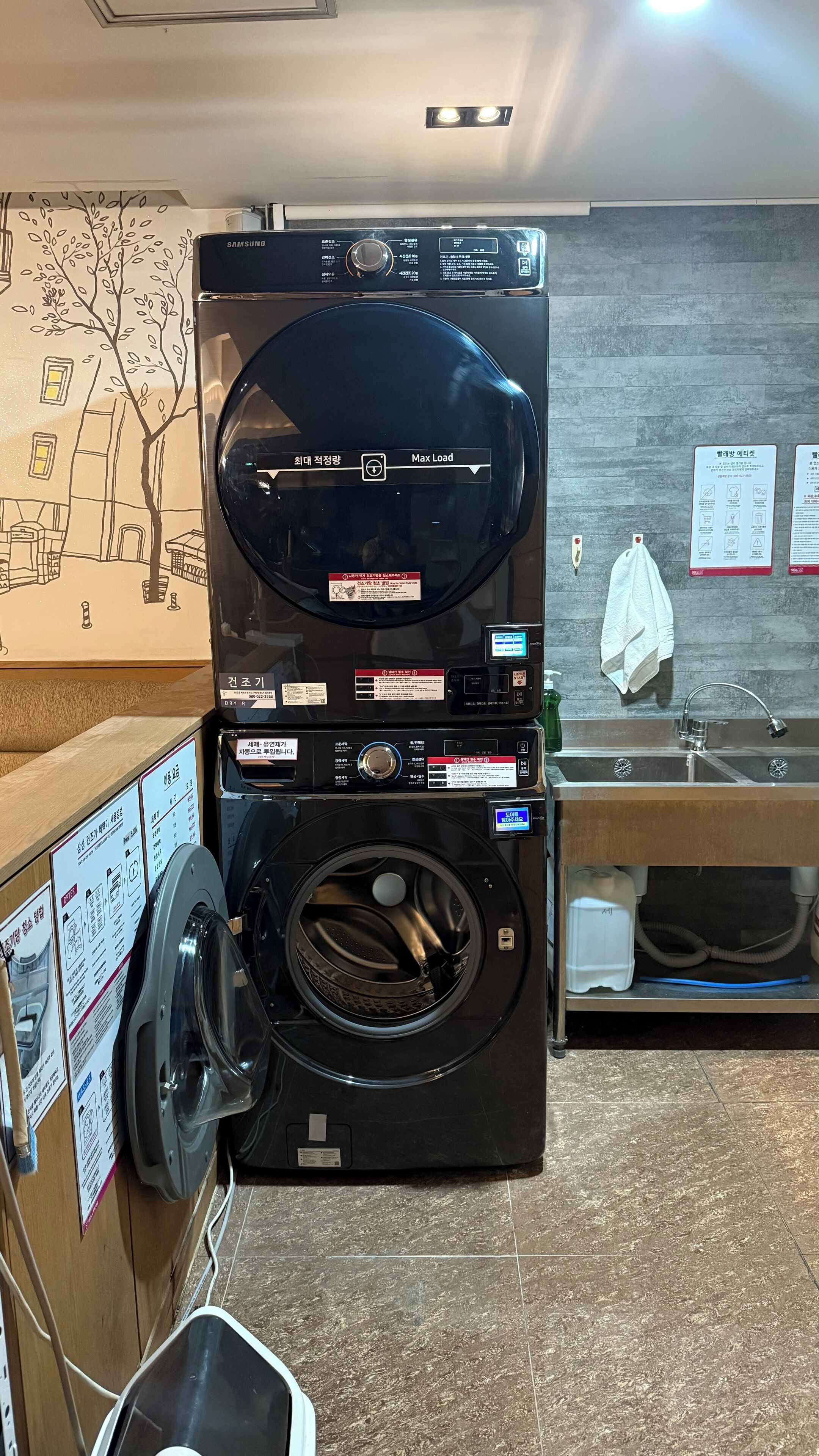 Eine moderne, kleine Waschküette mit einer Frontlader-Waschmaschine und einem Trockner. Deutliche Anweisungen und Sicherheitshinweise sind angebracht, ebenso ein kleines Waschbecken mit Handtuch und ein dekoratives Wandbild.