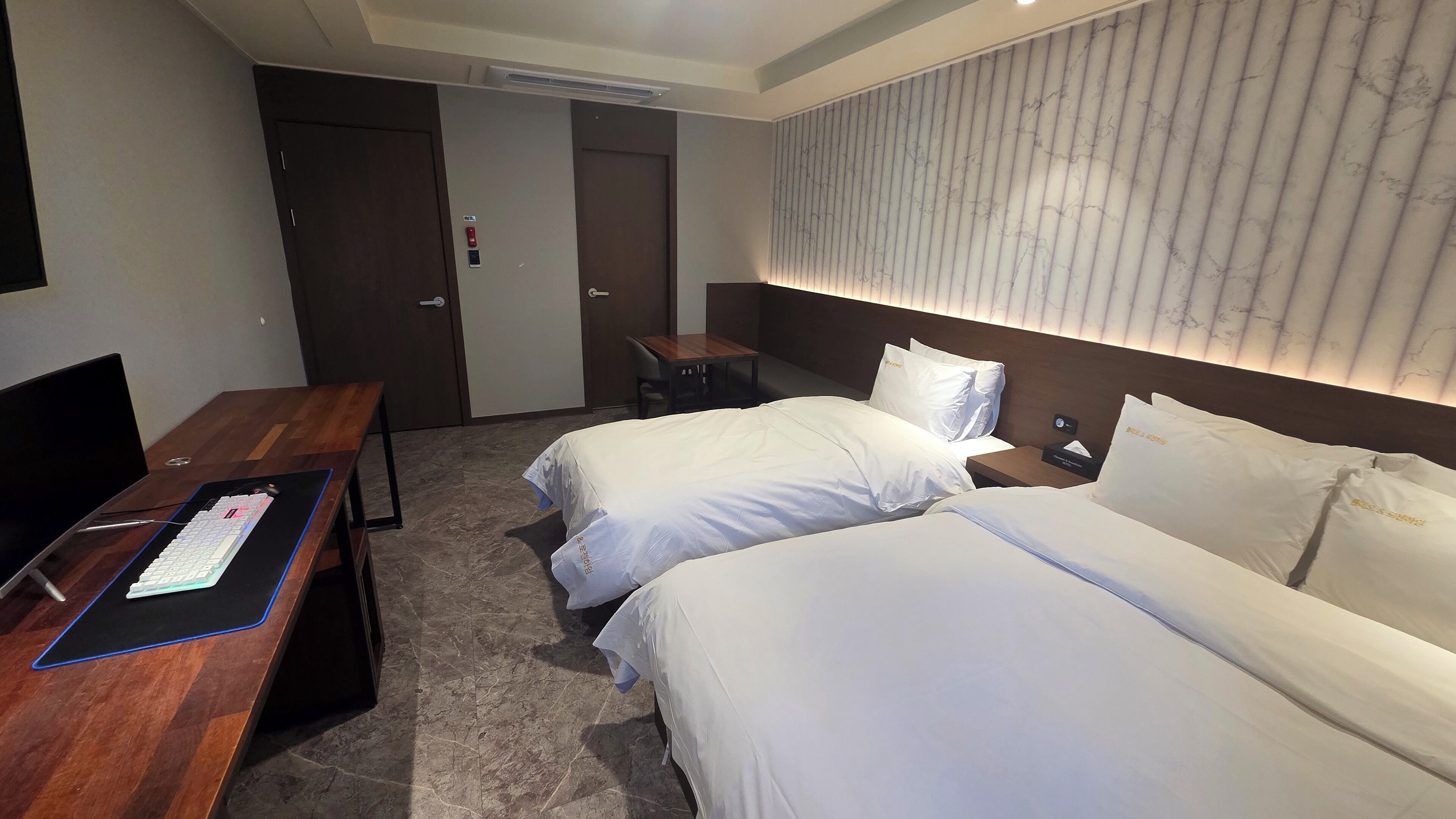 A hotel room with two beds, a desk with a computer, a small table, and a wall-mounted panel.
