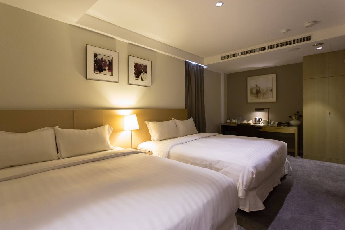 A hotel room with two beds, a desk with a lamp, a telephone, and a painting on the wall.