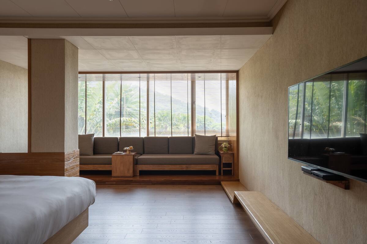 Une chambre moderne avec un grand lit, un canapé, un téléviseur fixé au mur et une fenêtre avec des stores.