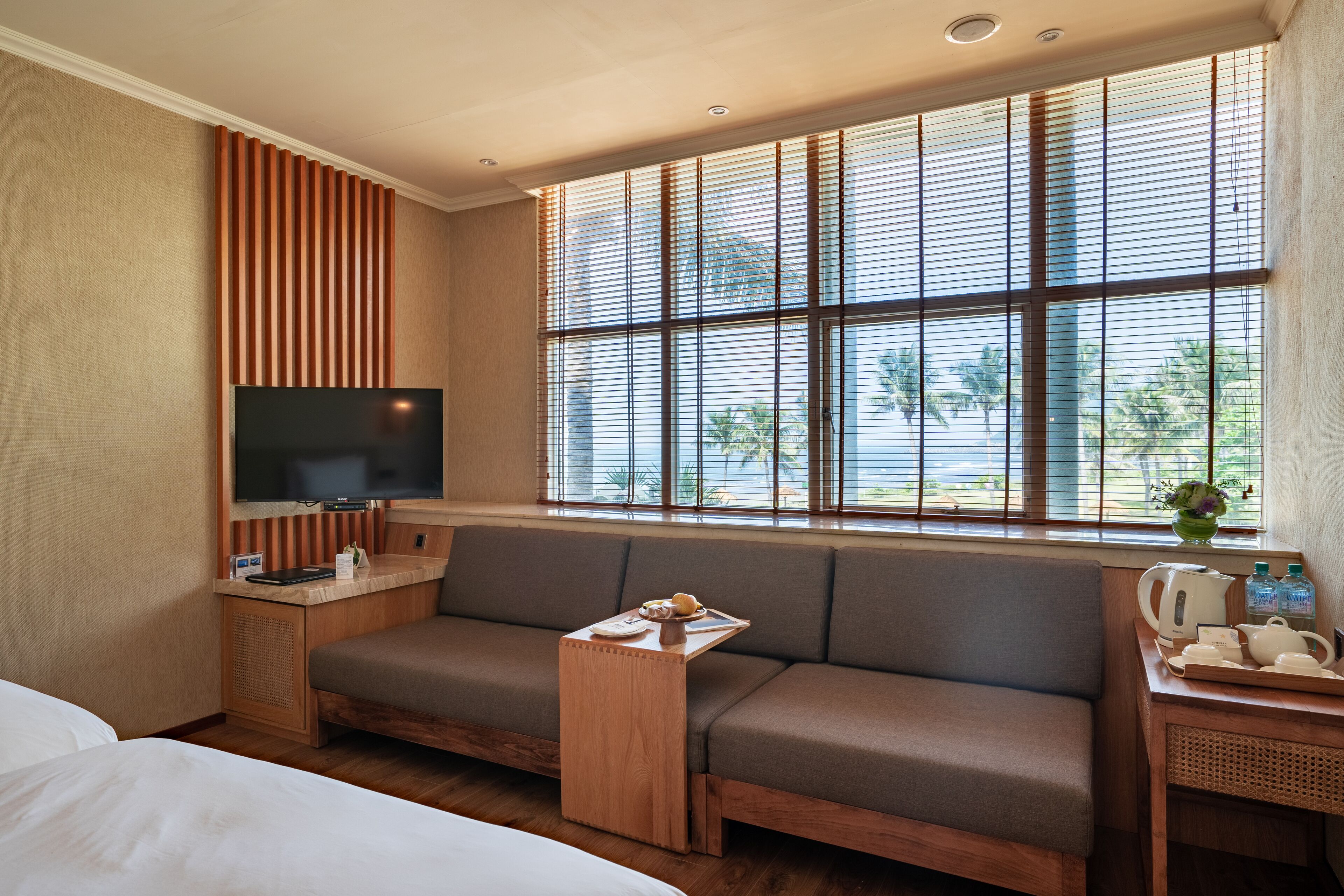 A hotel room with a large window, a sofa, a TV, and a wooden side table with a tray.