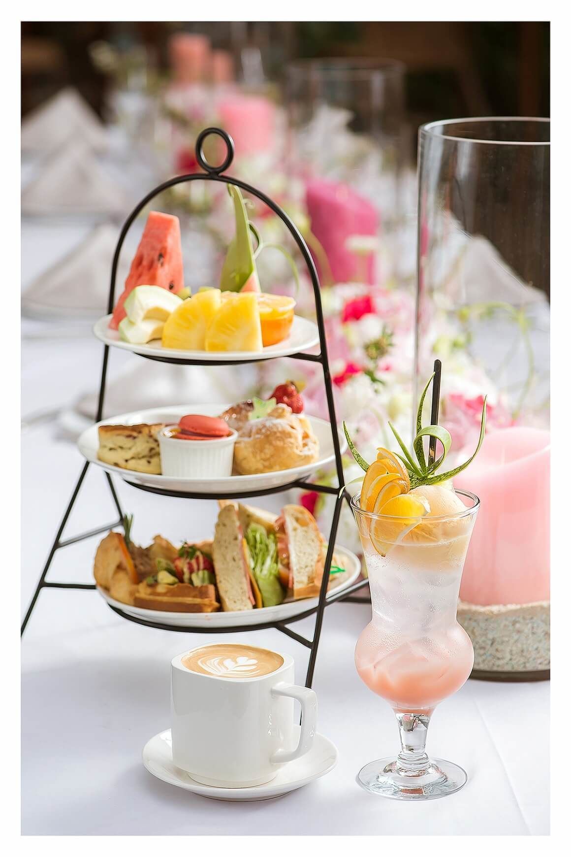 A tiered dessert stand with various pastries and fruits, a glass of iced drink with fruit garnish, and a cup of coffee on a table with a pink candle and flowers in the background.