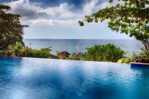 A clear view of a swimming pool with a view of the ocean and a tropical setting with trees and thatched-roof structures. - Amun Ini Beach Resort & Spa (Anda)