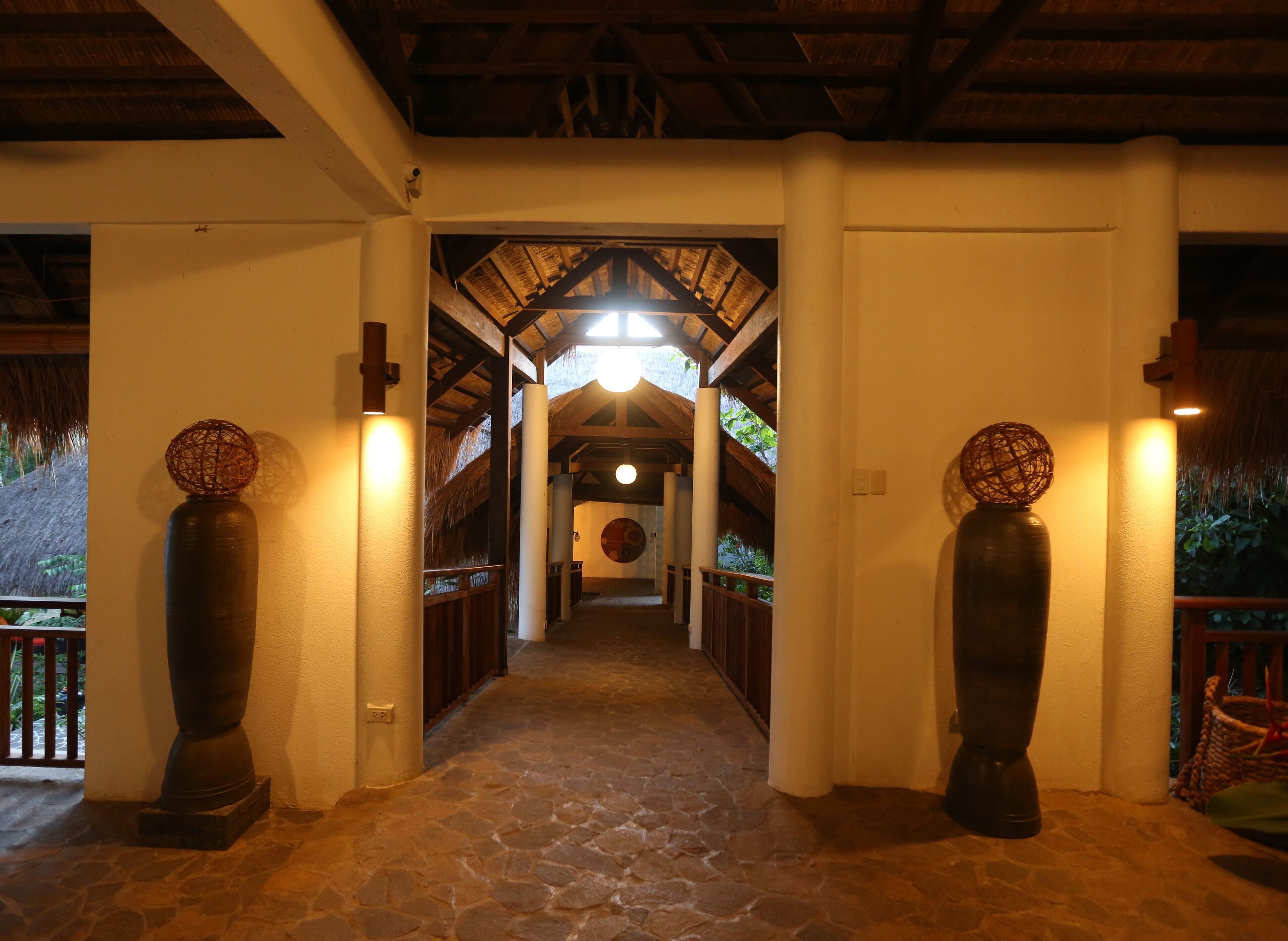 A stone-paved corridor with a thatched roof ceiling, leading to a circular room with a central light fixture.