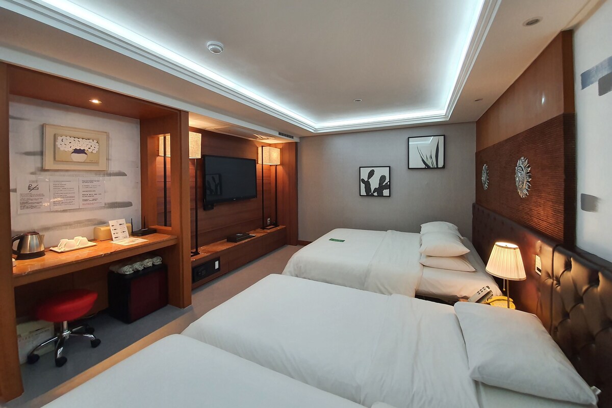 Une chambre d’hôtel équipée d’un bureau, de deux lits, d’une télévision et d’œuvres d’art accrochées aux murs.