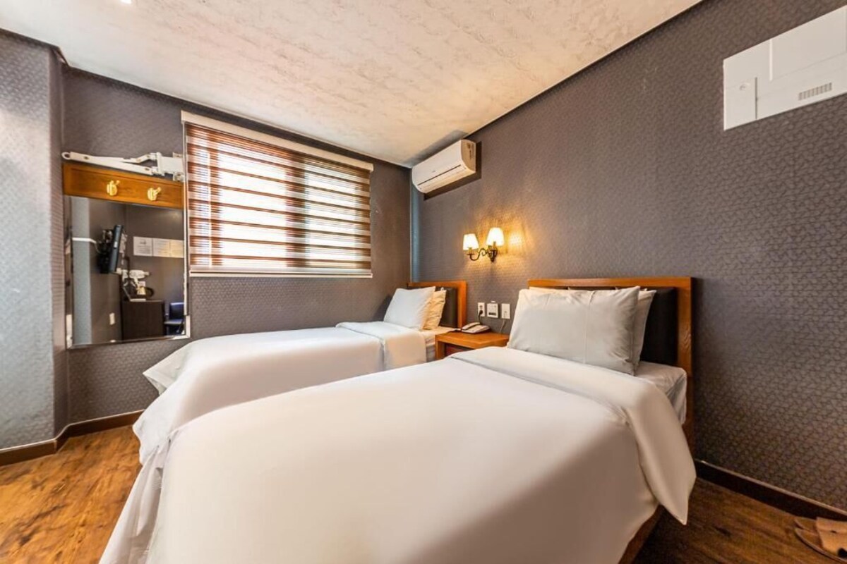 Une chambre d’hôtel avec deux lits, une grande fenêtre avec stores, un miroir et un climatiseur fixé au mur.