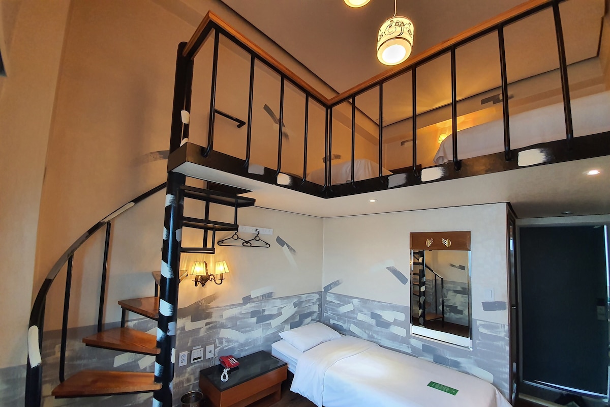 Une chambre d’hôtel moderne avec un espace nuit en mezzanine, une petite table de chevet, une lampe et une fenêtre donnant sur un paysage agréable.