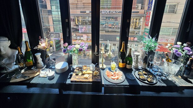 A table set with various food items and drinks, viewed through a window with a cityscape in the background. - Friendly DH Naissance Hotel by Mindrum Group (Seoul)