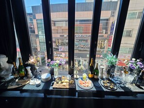 Una mesa servida con diversos alimentos y bebidas, vista a través de una ventana con un paisaje urbano de fondo.