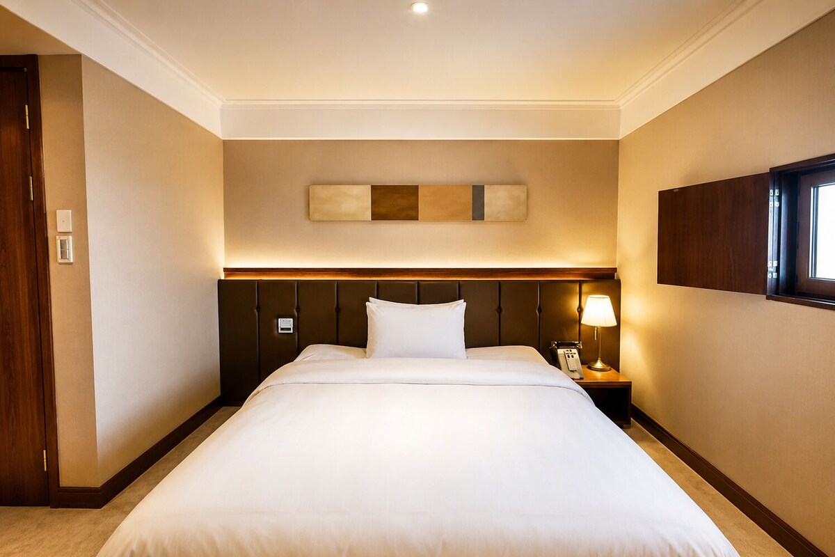 Une chambre d’hôtel avec un grand lit, une tête de lit ornée de lignes verticales, une table de chevet avec une lampe et un téléviseur fixé au mur.