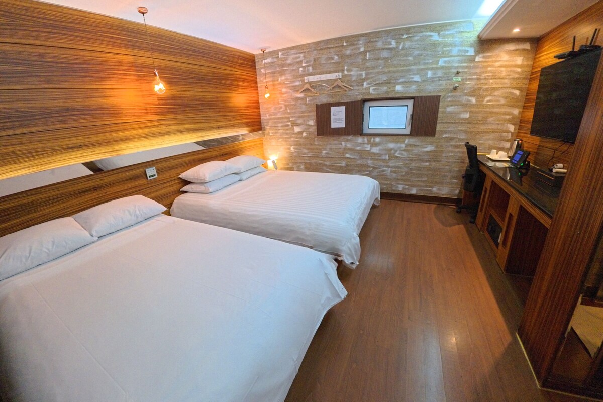 Une chambre d’hôtel avec deux lits, des têtes de lit en bois, un mur décoratif en pierre, une télévision et un bureau avec un ordinateur.
