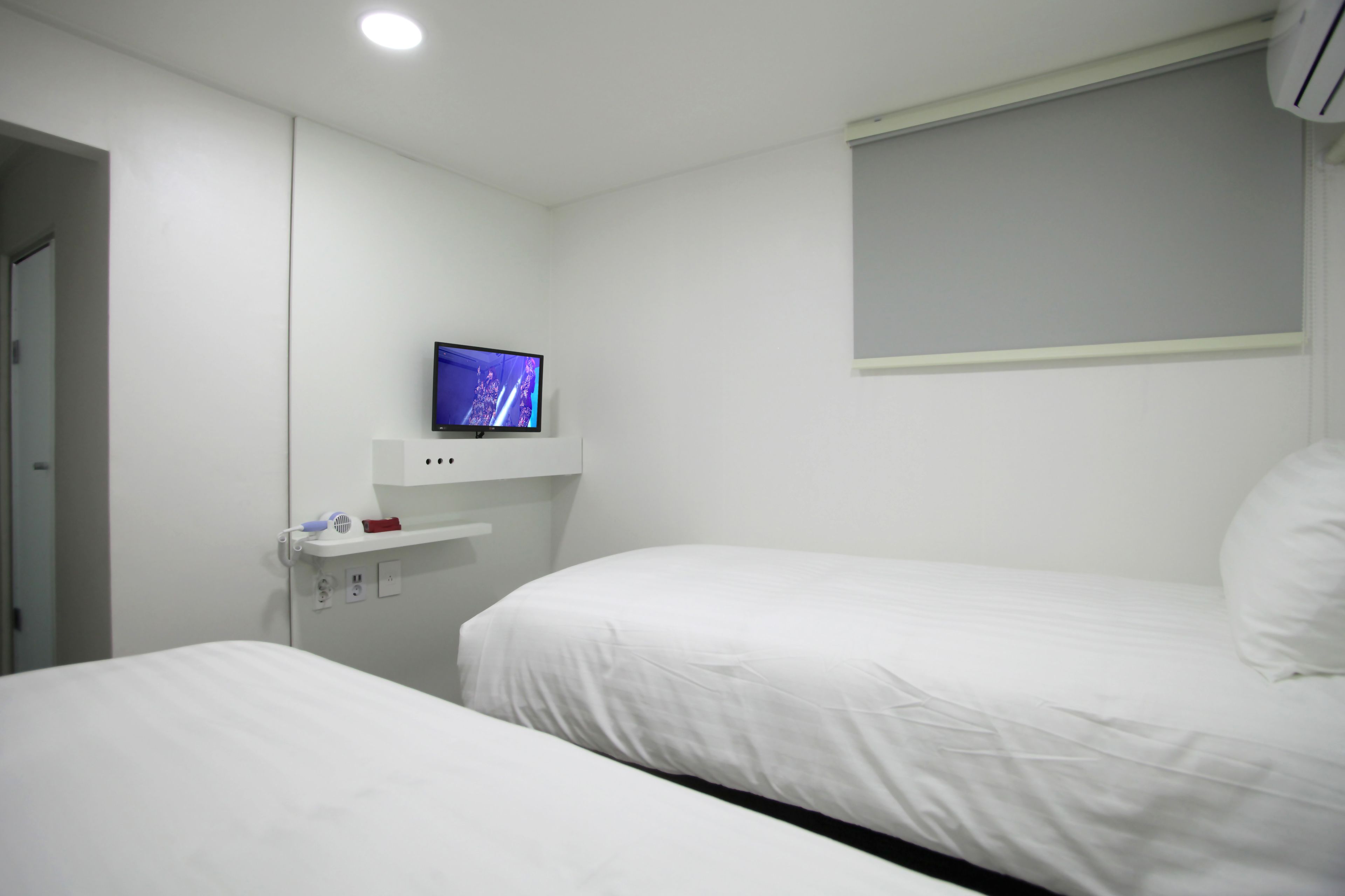 A hotel room with two single beds, a flat-screen TV mounted on the wall, and a small shelf with a phone and a red object.