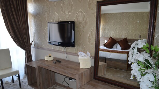 A hotel room with a wooden desk, a flat-screen TV mounted on the wall, a large mirror, and a bed with white bedding and brown pillows. - Nobel Hotel Ankara (Ankara)