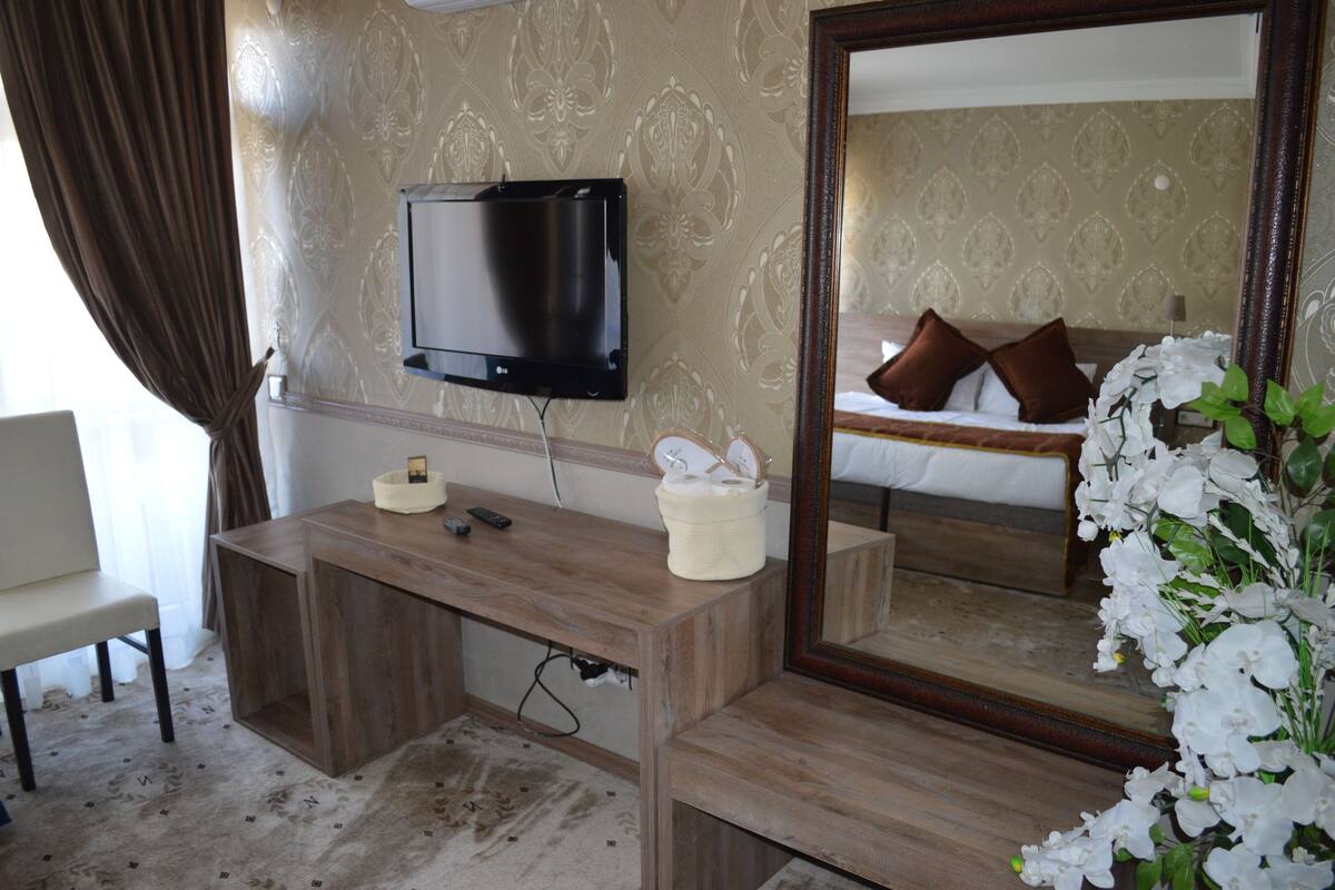 A hotel room with a wooden desk, a flat-screen TV mounted on the wall, a large mirror, and a bed with white bedding and brown pillows.