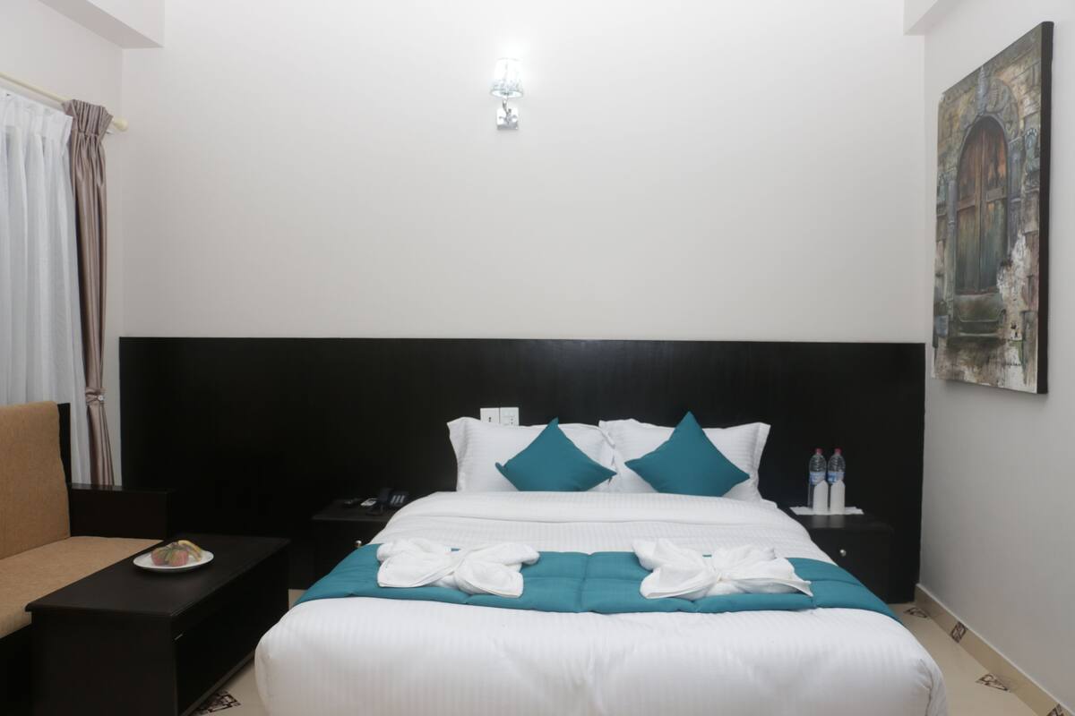 A hotel room with a large bed, a nightstand, a bedside table, a chair, and a painting on the wall.