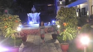Un espacio exterior nocturno con una fuente, plantas en macetas y un muro de ladrillo.