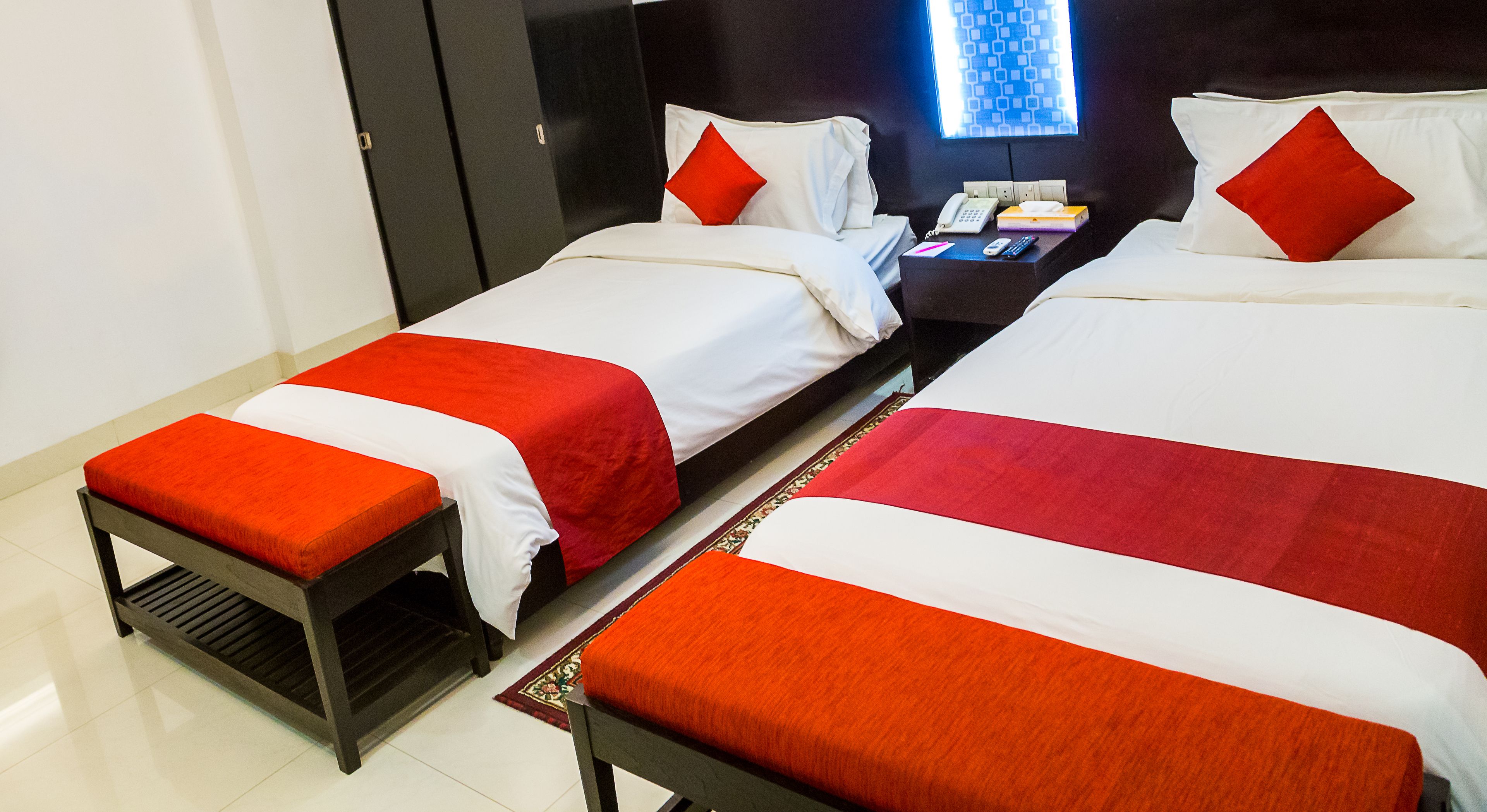 A hotel room with two beds, each with white and red bedding, a small table with a phone and remote controls, and a window with a geometric pattern.