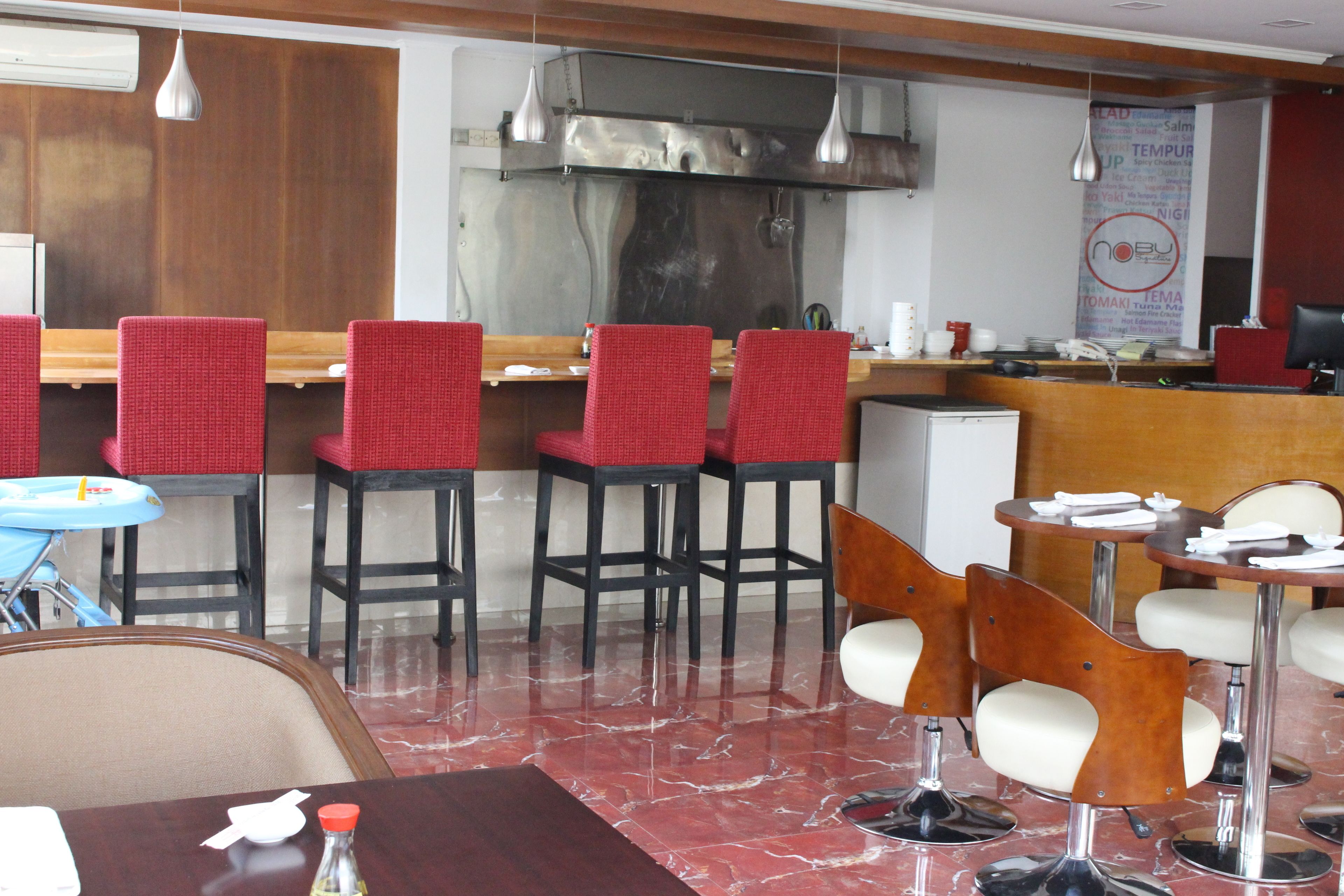 A restaurant interior with red upholstered chairs, a marble floor, and a stainless steel kitchen in the background.