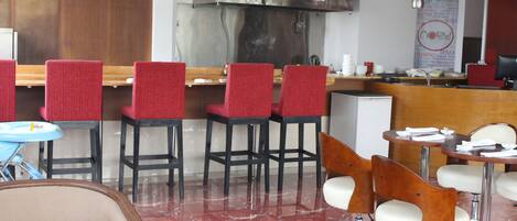 A restaurant interior with red upholstered chairs, a marble floor, and a stainless steel kitchen in the background.