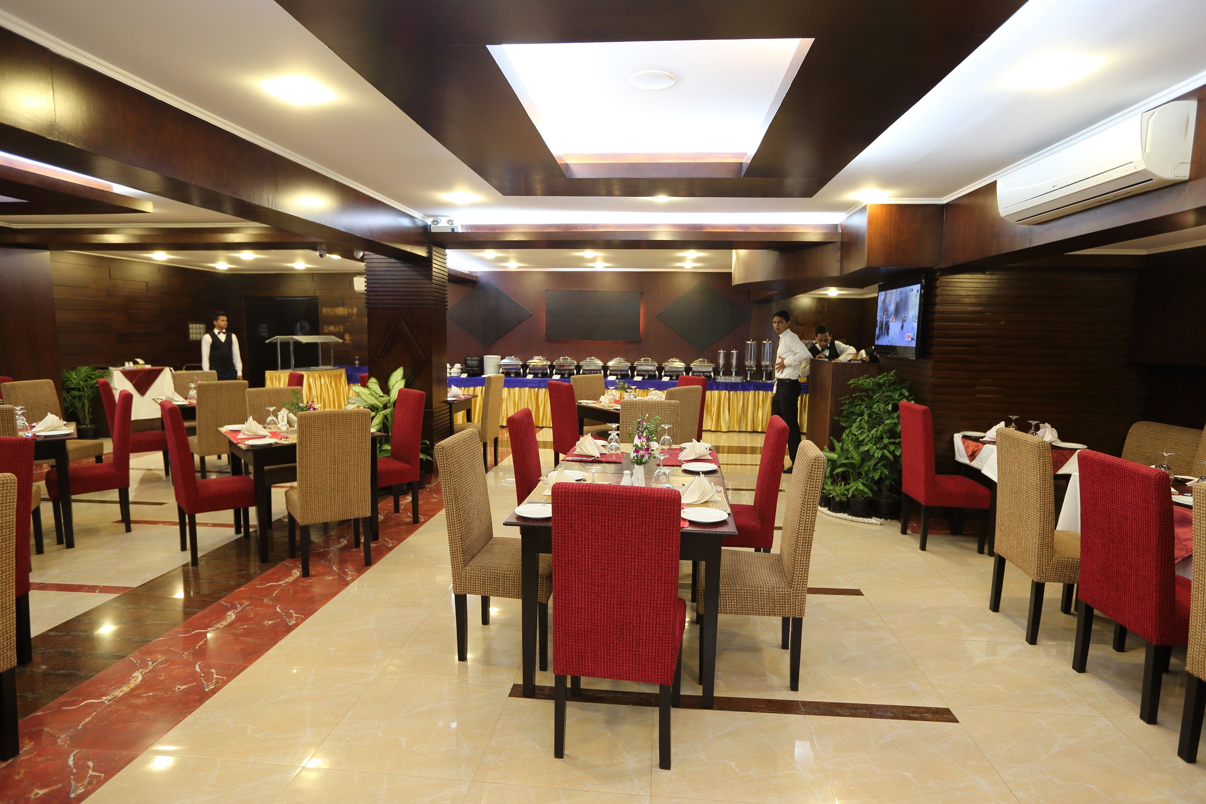 A hotel dining area with multiple tables and chairs, a stage with a podium, and a television screen.