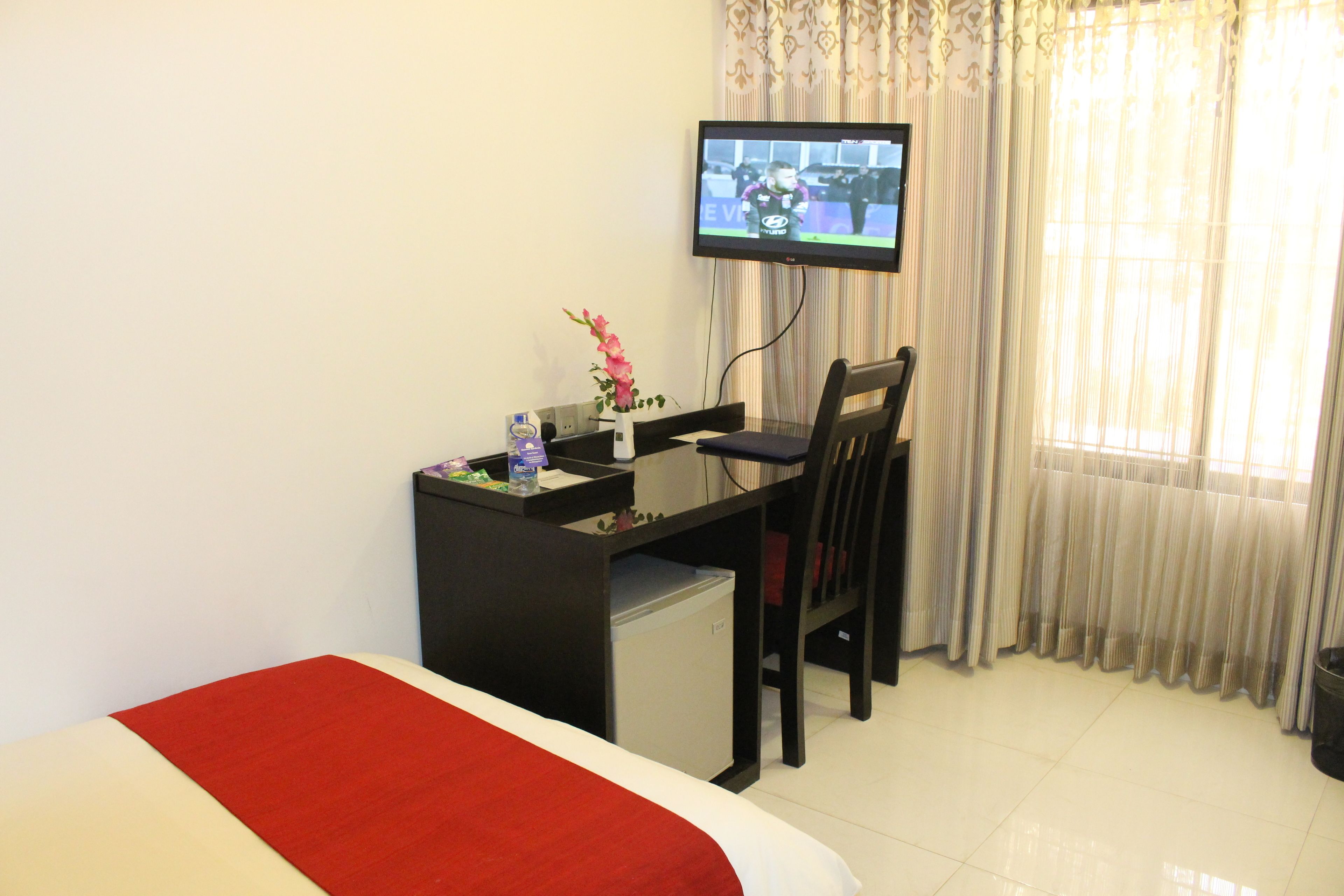 A hotel room with a bed, a desk with a computer, a chair, a TV mounted on the wall, and a window with curtains.