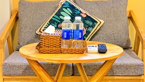 A round wooden table with a woven basket, two water bottles, and a remote control on a grey sofa with a floral cushion.