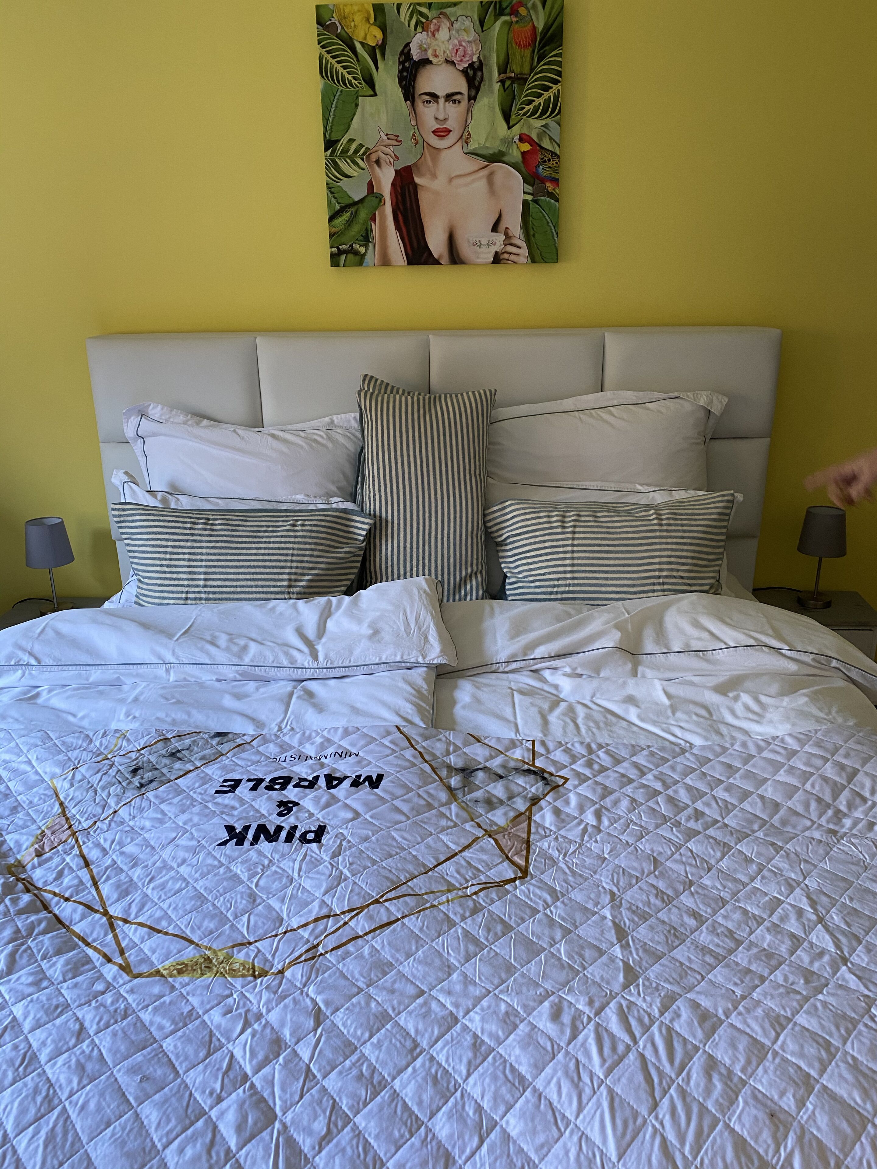 A bed with a quilted white comforter, multiple pillows, and a wall art piece above.