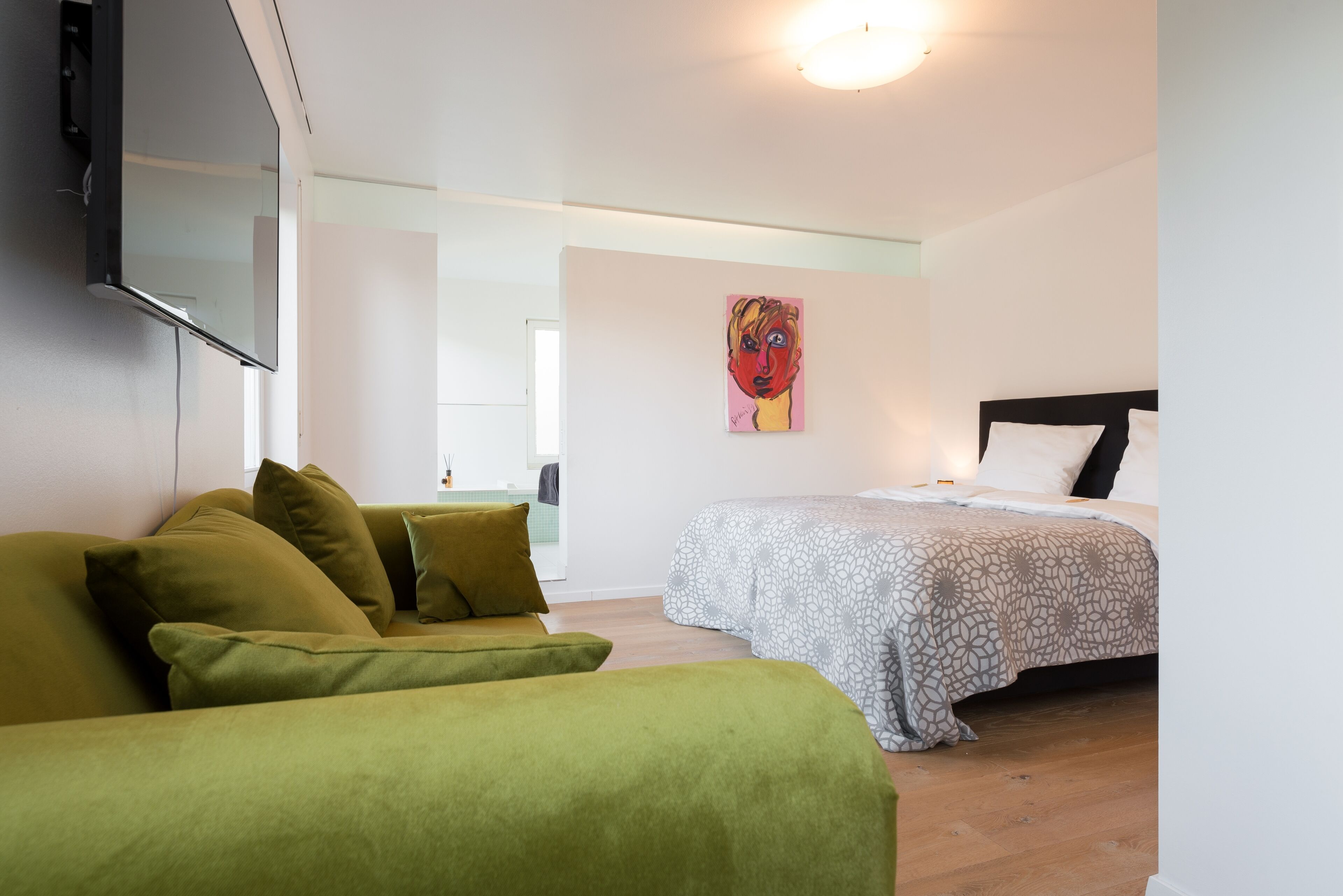 A room with a bed, a green sofa, a painting on the wall, and a flat-screen TV mounted on the wall.