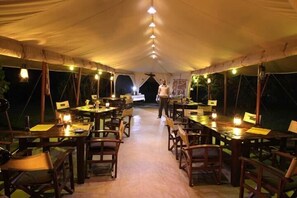 A tented dining area with tables and chairs, a central walkway, and hanging lights. - Mara Bush Camp (Maasai Mara)