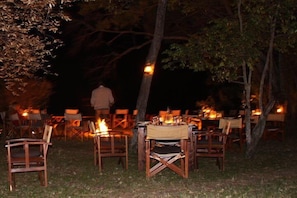 An outdoor dining area with tables and chairs arranged under trees, illuminated by candlelight. - Mara Bush Camp (Maasai Mara)