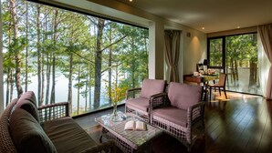 A spacious room with a large window offering a view of a forest and lake. There is a wicker sofa, a small table with a book, and a chair. - Terracotta Hotel & Resort Dalat (Xuan Huong - Da Lat)