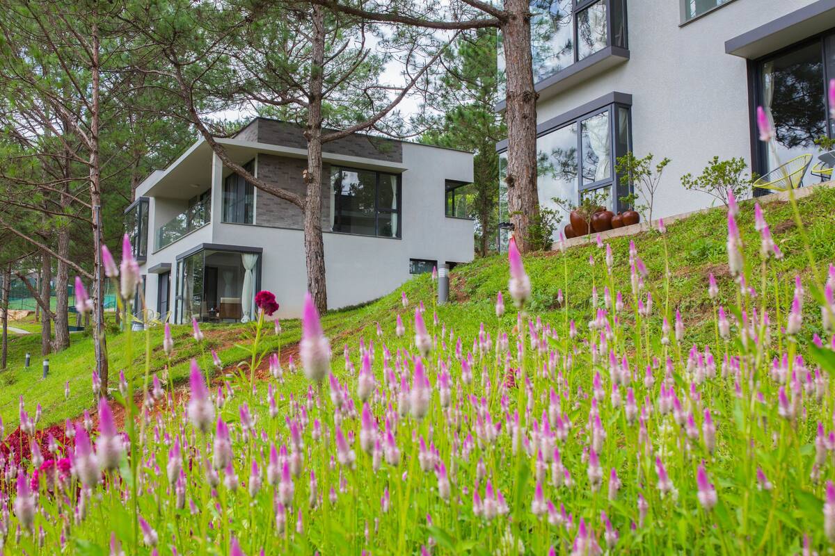 Ein modernes, zweistöckiges Haus mit Balkon, umgeben von üppigem Grün und blühenden Pflanzen.