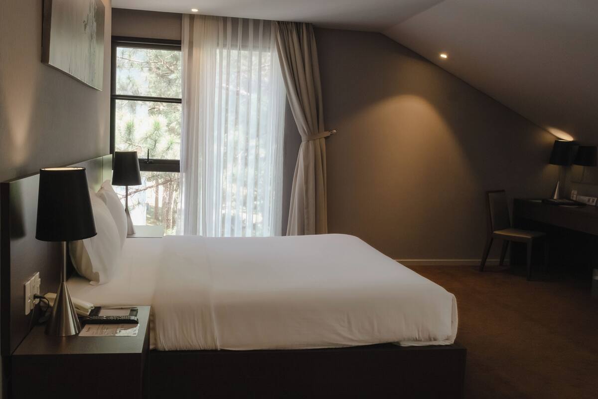 Ein modernes Hotelzimmer mit einem großen Bett, einem Schreibtisch mit Lampe und Blick auf Bäume durch das Fenster.