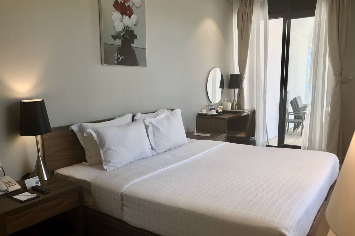 Ein Hotelzimmer mit einem großen Bett, einem Nachttisch mit Lampe, einem Schreibtisch mit Spiegel und einem Fenster mit Vorhängen.