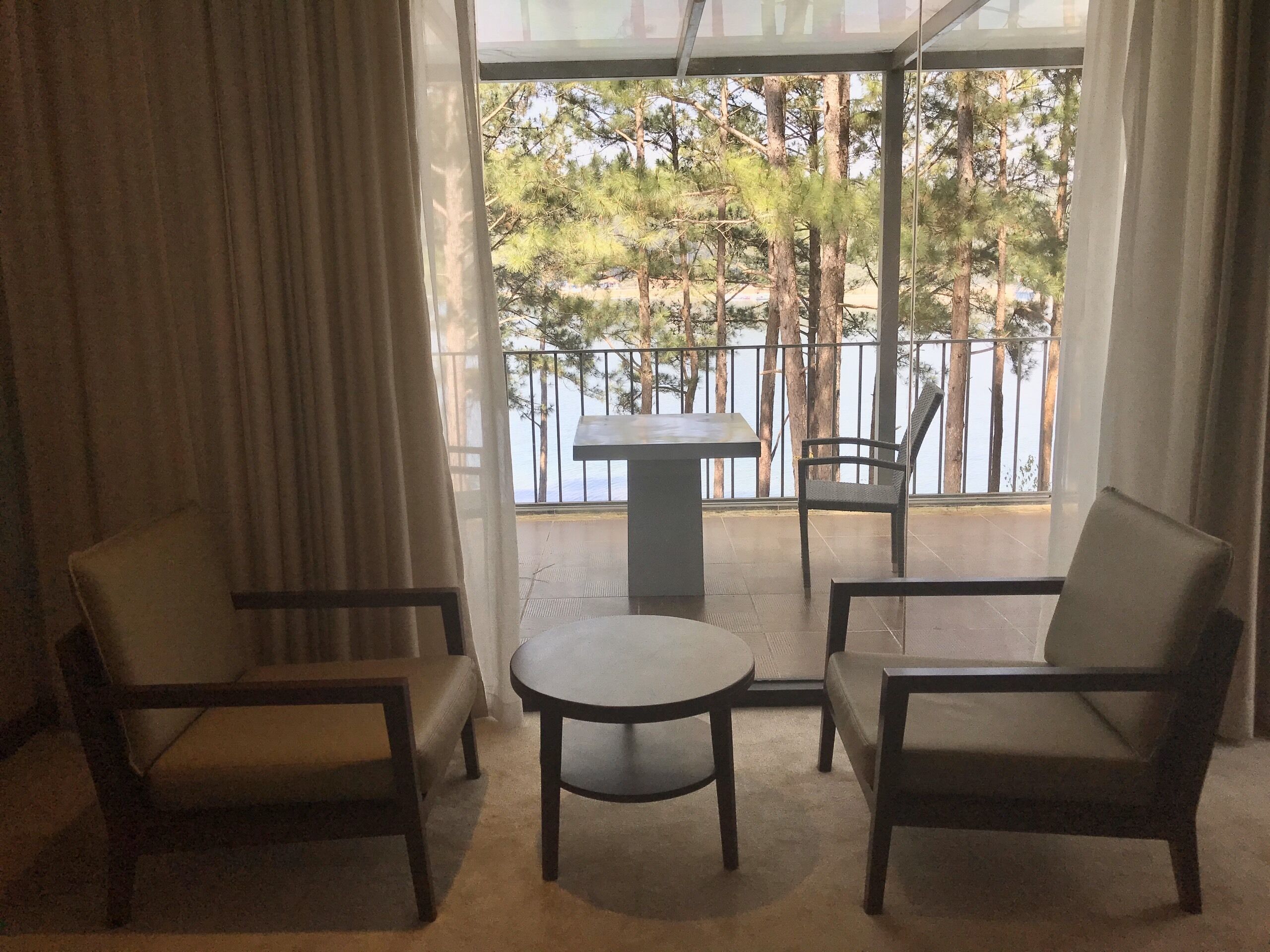 A room with two chairs and a small table, a view of trees and a railing through a window.