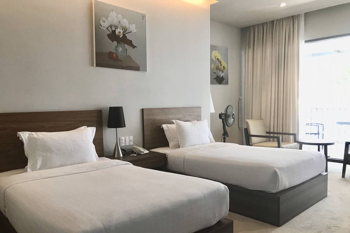 Ein Hotelzimmer mit zwei Betten, einem Nachttisch mit Lampe, einem Sessel und Bildern an der Wand.