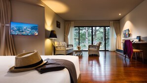 A hotel room with a bed, a TV, a desk, wicker chairs, and a large window. - Terracotta Hotel & Resort Dalat (Xuan Huong - Da Lat)