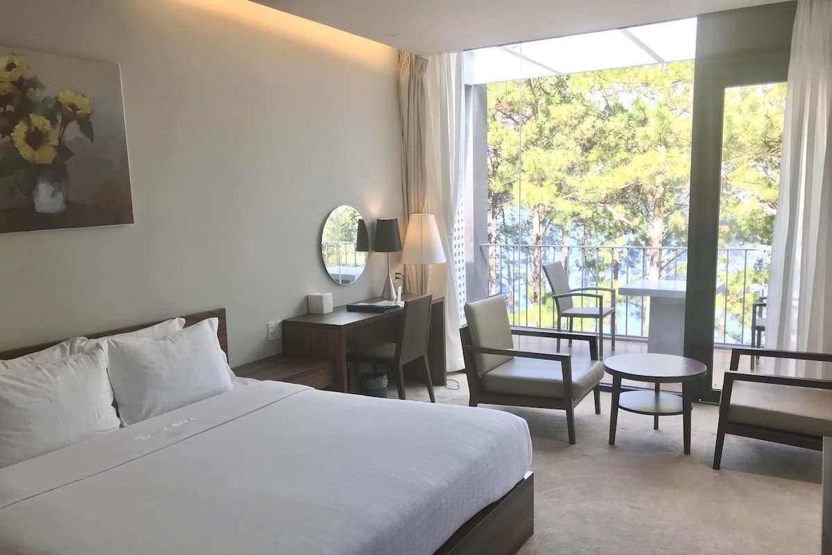 Ein Hotelzimmer mit einem großen Bett, einem Schreibtisch mit Stuhl, einem kleinen Tisch und einem Balkon mit Blick auf Bäume.