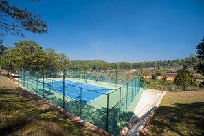 Un court de tennis en plein air, entouré d’une clôture verte, avec un ciel bleu dégagé au-dessus et des arbres en arrière-plan.