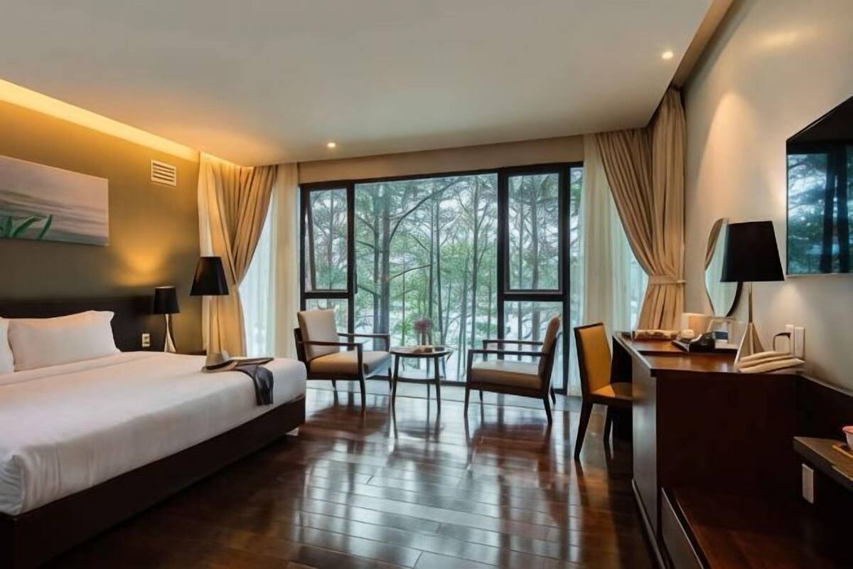 Ein Hotelzimmer mit einem großen Bett, zwei Stühlen, einem kleinen Tisch, einem Schreibtisch und einem großen Fenster mit Blick auf Bäume.