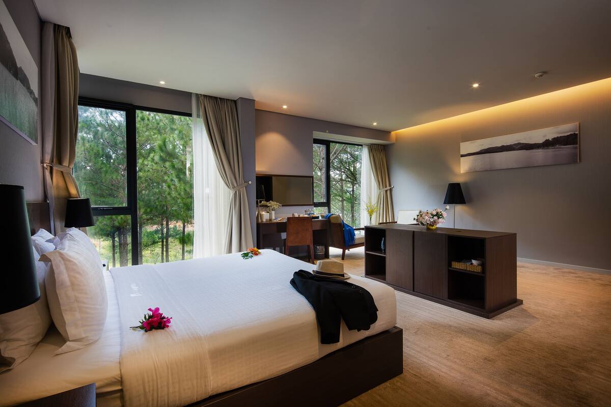 Ein modernes Hotelzimmer mit einem großen Bett, einem Schreibtisch und Blick auf Bäume durch große Fenster.